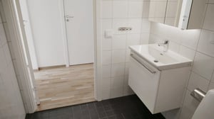 Ljust badrum med vita kakelväggar, vit kommod med spegel och svart klinkergolv, samt en öppning mot ett rum med parkettgolv