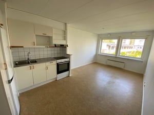 Kök och vardagsrum i en lägenhet med vita väggar, ljusbrunt golv, och fönster som vetter mot en innergård. Köksdelen har vita skåp, vit kyl och spis med svart häll samt kakel som stänkskydd