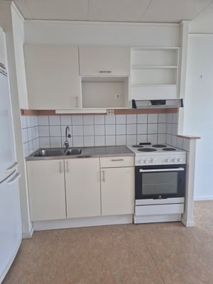 Litet kök med vita skåp, diskho, spis och ugn. Vägg med kakel ovanför köksbänken
