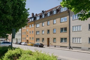 Gul flerfamiljsbyggnad med tre våningar och bruna takpannor, flera fönster och parkerade bilar framför