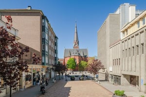 Flerbostadshus i tegel med gröna burspråk, butiker på bottenvåningen och trottoarer längs en gågata med promenader