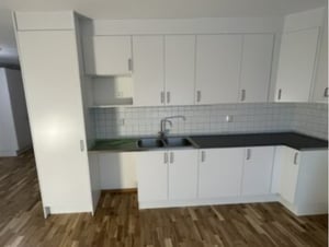 Vit köksinredning med flera skåp, diskho, grå bänkskivor och kaklat stänkskydd. Golvet är av trä och rummet har naturligt ljus