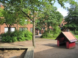 Lekplats med sand, ett rött lekhus och träbänkar, omgiven av grönskande träd och buskar framför röda tegelbyggnader