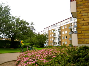 Lägenhetsbyggnad med gul fasad omgiven av grönska och rosa blommor, med en lekplats och gångväg i förgrunden