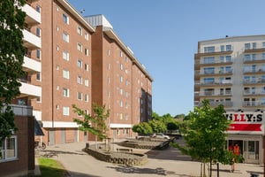 Bostadshus i brun fasad med flera våningar med balkonger, omgiven av parkering och handel under en klar himmel
