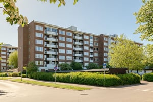 Bostadshus i brun fasad med flera våningar med balkonger, omgiven av grönska på innergård under en klar himmel