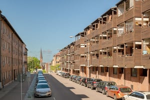 Flerbostadshus i rött tegel med bruna balkonger, med parkerade bilar längs lokalgata