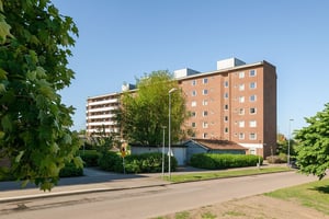 Bostadshus i brun fasad med flera våningar med balkonger, omgiven av grönområde under en klar himmel