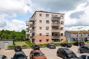 Flerfamiljshus med tegelfasad, balkonger omgiven av grönska och parkeringar framför huset under klar himmel