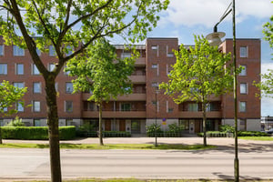 Bostadshus i tegel med flera våningar med balkonger, omgiven av gata och träd under en molnig himmel
