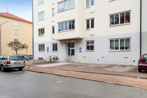 Flerfamiljshus med vit fasad, flera fönster och entré. Parkering och cyklar synliga framför byggnaden
