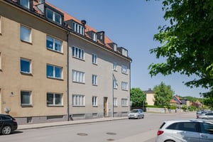 Flerfamiljshus med beige fasad, parkerade bilar längs gatan och ett gult hus i bakgrunden under en klar himmel