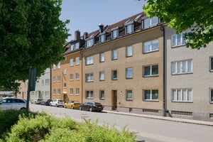 Gul flerfamiljsbyggnad med tre våningar och bruna takpannor, flera fönster och parkerade bilar framför