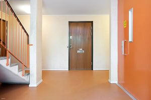 Entréområde med en trappa till vänster, en brun dörr i mitten och en orange dörr till höger. Väggarna är ljusa och golvet är i orange ton