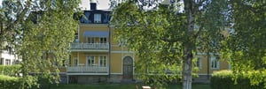 Gult flerfamiljshus med vita balkonger och stora träd framför