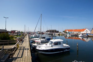 Båtar förtöjda vid en liten hamn med brygga, omgivna av byggnader och klarblå himmel