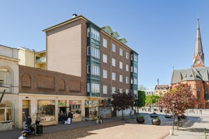 Flerbostadshus i tegel med gröna burspråk, butiker på bottenvåningen och trottoarer längs en gata med promenader och trafik