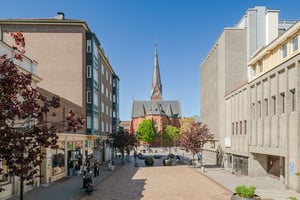 Flerbostadshus i tegel med gröna burspråk, butiker på bottenvåningen och trottoarer längs en gågata med promenader