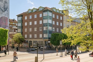 Flerbostadshus i tegel med gröna burspråk, butiker på bottenvåningen och trottoarer längs en gata med promenader och trafik