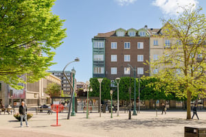 Flerbostadshus i tegel med gröna burspråk, omgiven av torg med folk i rörelse