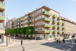 Flerbostadshus i tegel med gröna balkonger, butiker på bottenvåningen och trottoarer längs en gata med promenader och trafik
