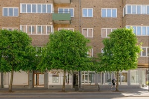 Flerbostadshus i tegel med gröna balkonger, butiker på bottenvåningen och trottoarer längs en gata med promenader och trafik