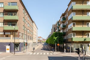 Flerbostadshus i tegel med gröna balkonger, butiker på bottenvåningen och trottoarer längs en gata med promenader och trafik