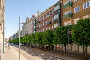 Flerbostadshus i tegel med gröna burspråk, butiker på bottenvåningen och trottoarer längs en gata med promenader och trafik