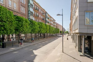Flerbostadshus i tegel med gröna burspråk, butiker på bottenvåningen och trottoarer längs en gata med promenader och trafik