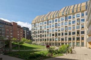 Flerbostadshus i ljus fasad med balkonger mot stora fönster mot innergård med grönska och lekplats