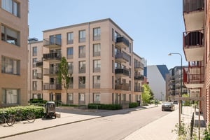 Flerfamiljshus i ljust tegel med balkonger omgivet av grönska och en gata med parkeringar och cyklar