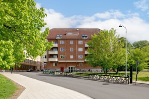 Flerfamiljshus i tegelfasad med takkupor och vita balkonger, omgiven av gräsmatta, träd och en gata med cykelställ i förgrunden