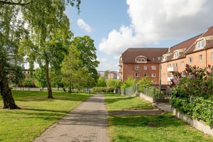 Flerfamiljshus i tegelfasad med takkupor och vita balkonger, omgiven av gräsmatta, träd och en stenbelagd gångbana