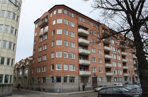Röd tegelbyggnad med flera våningar, balkonger och stora fönster, omgiven av parkerade bilar och träd