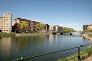 Flerfamiljshus på avstånd, i tegel och ljusa balkonger, omgiven av grönska och gata samt vattenkanal i förgrunden