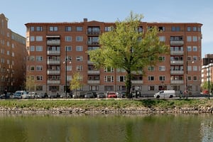 Flerfamiljshus i tegel och ljusa balkonger, omgiven av grönska och gata med vattenkanal i förgrunden