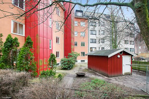 Bostadsgård med rödmålat skjul, buskar och träd, omgiven av flerbostadshus i orange och vita fasader