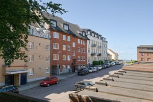 Flerbostadshus i rött tegel med svart tak, omgiven av andra fastigheter i ljusa fasader och gata med parkerade bilar