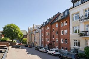 Flerbostadshus i rött tegel med svart tak, omgiven av andra fastigheter i ljusa fasader och gata med parkerade bilar