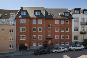 Flerbostadshus i rött tegel med svart tak, med infart till innergård till vänster i fasaden, parkerade bilar framför huset