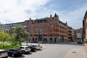 Flerbostadshus i rött tegel och grå puts med detaljer kring fönster och torn på gavel, längs gata med parkerade bildar framför huset och vid parkeringsplats