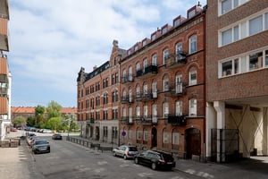 Flerbostadshus i rött tegel och grå puts med detaljer kring fönster och torn på gavel, längs gata med parkerade bilar
