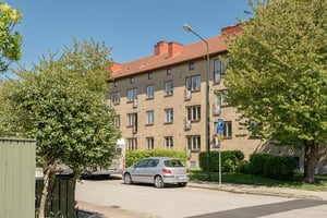 Flerfamiljshus i tegel med franska balkonger, omgiven av grönska och gata med en grå bil parkerad i förgrunden