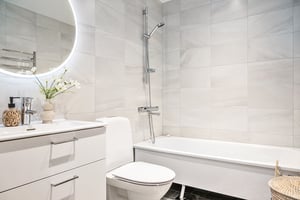 Modernt badrum med vita kakelväggar, rund spegel med belysning, toalett, badkar och handfat med dekorativ vas och tvålpump