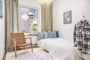 Sovrum med enkelbädd, ljusa väggar, vävstol, blå dekorativa kuddar och en bricka med blommor på fönsterbrädan. En gråvit rutmönstrad skjorta hänger på en krok