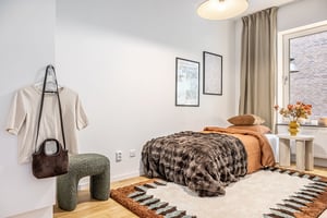 Sovrum med en enkelsäng klädd i bruna och beige sängkläder, en matta med geometriskt mönster, vägghängd konst, och ett fönster med beige gardiner