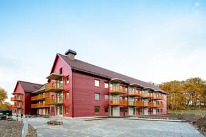 Röd flerfamiljsbyggnad med träfasad, flera balkonger och omgivande grönska under en klar himmel