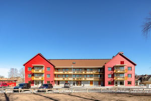 Rött flerbostadshus med balkonger och parkerade bilar framför under klarblå himmel