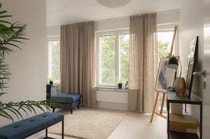 Inflyttningsklar lägenhet med ljust vardagsrumsmiljö, stora fönster, beige gardiner och blå sittbänkar