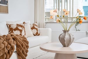 Ljus vardagsrum med vit soffa, bruna mönstrade kuddar, beige filt och blomvas med orange och vita blommor på ett runt träbord